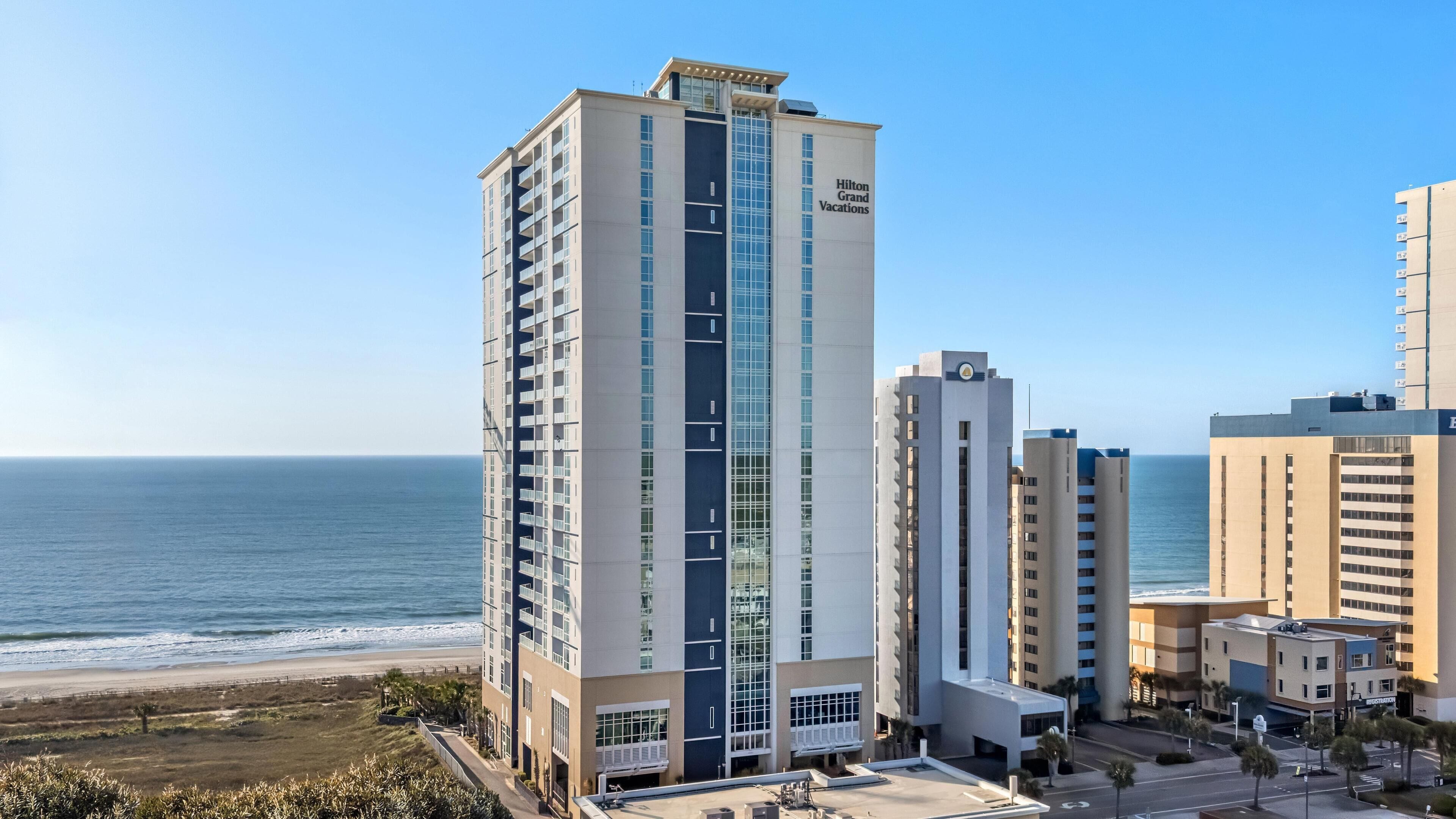 A high-rise hotel with a prominent glass facade, located near a beach with a clear view of the ocean.