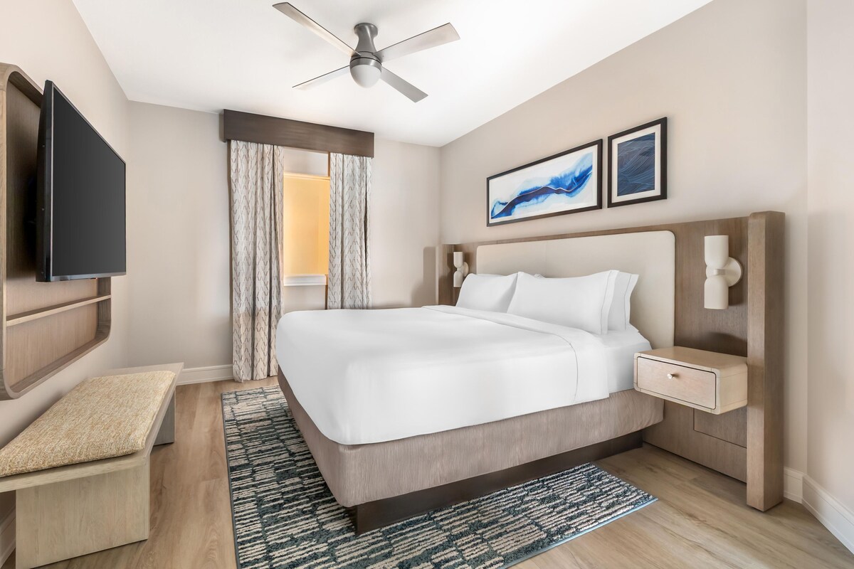 Habitación de hotel con cama, mesitas de noche, televisor de pantalla plana, ventilador de techo y obras de arte en la pared.