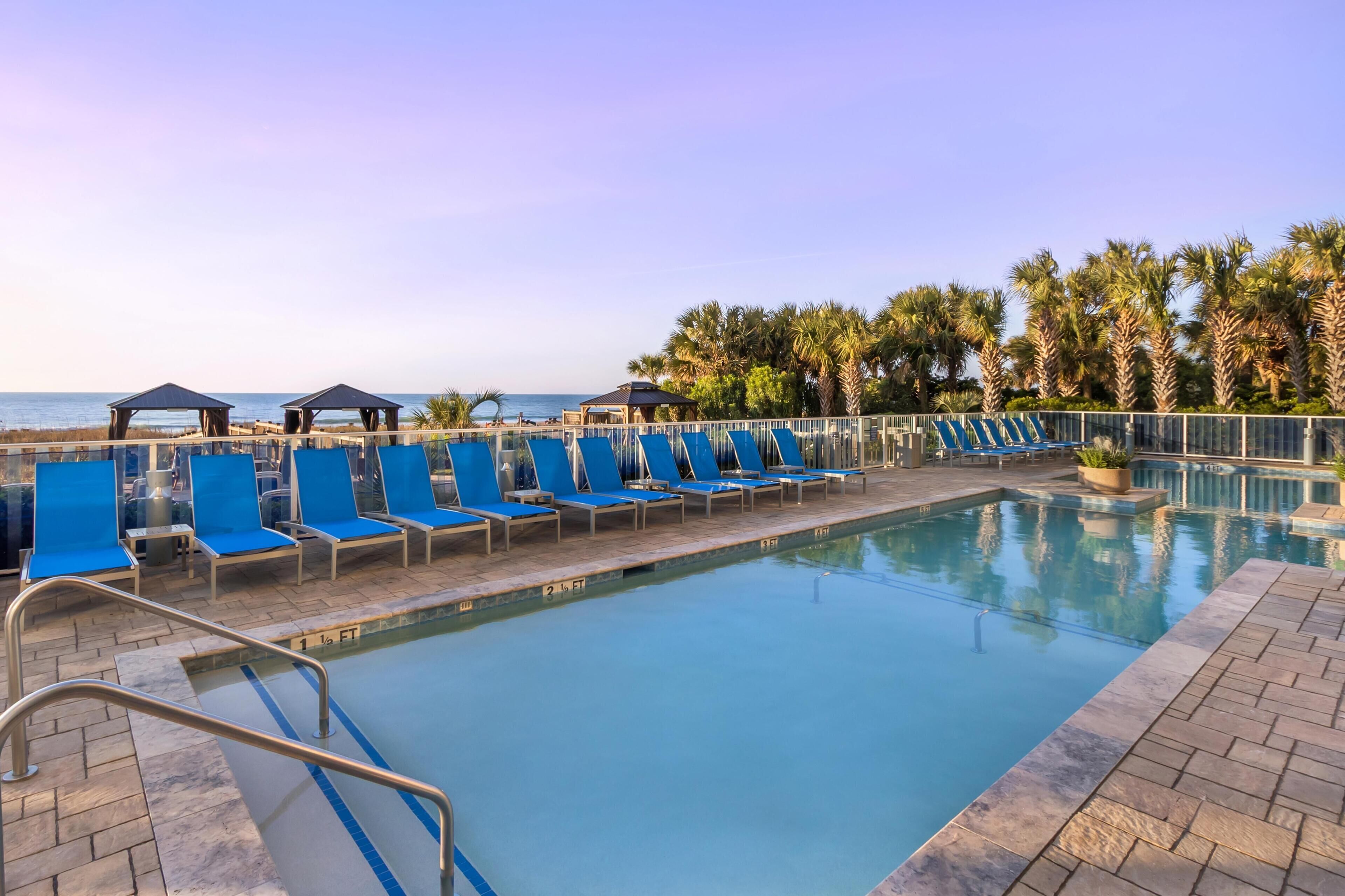 Un área de piscina con sillones reclinables azules, vista despejada del mar y palmeras.