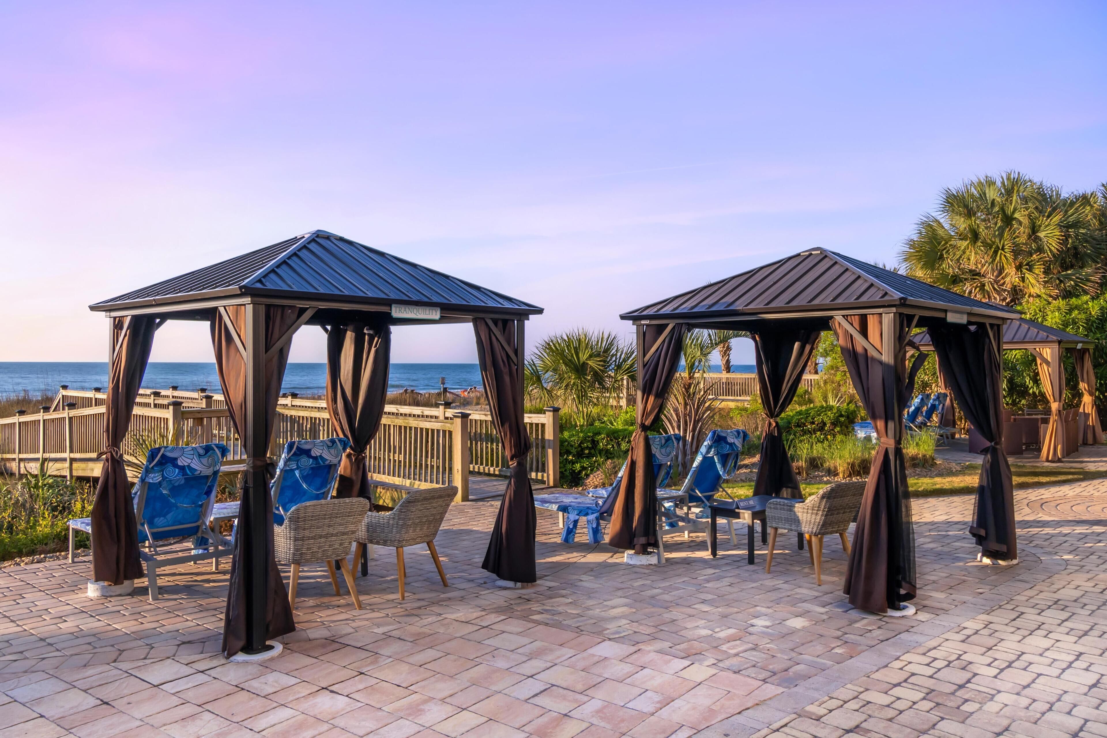 Dos gazebos de madera con sillas azules y muebles de mimbre, una terraza de madera y vista al océano.