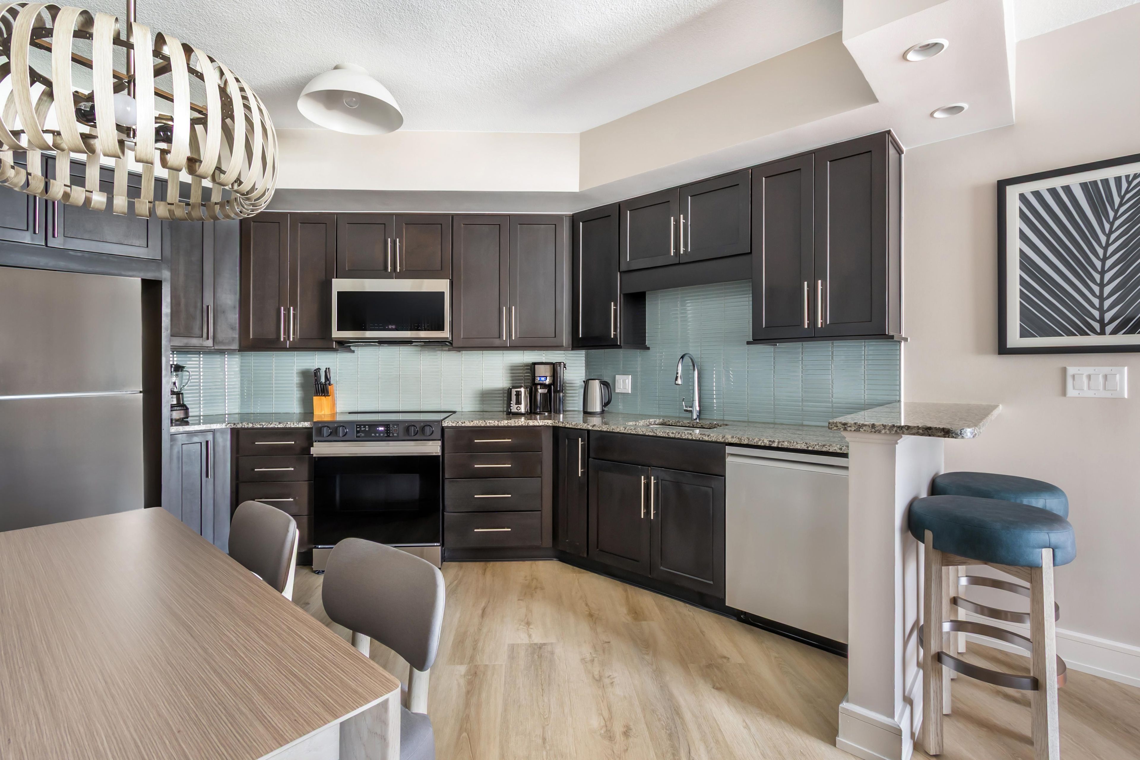 A modern kitchen with dark cabinets, a stainless steel refrigerator, a microwave, and a coffee maker. There is a wooden dining table with chairs and a bar stool.