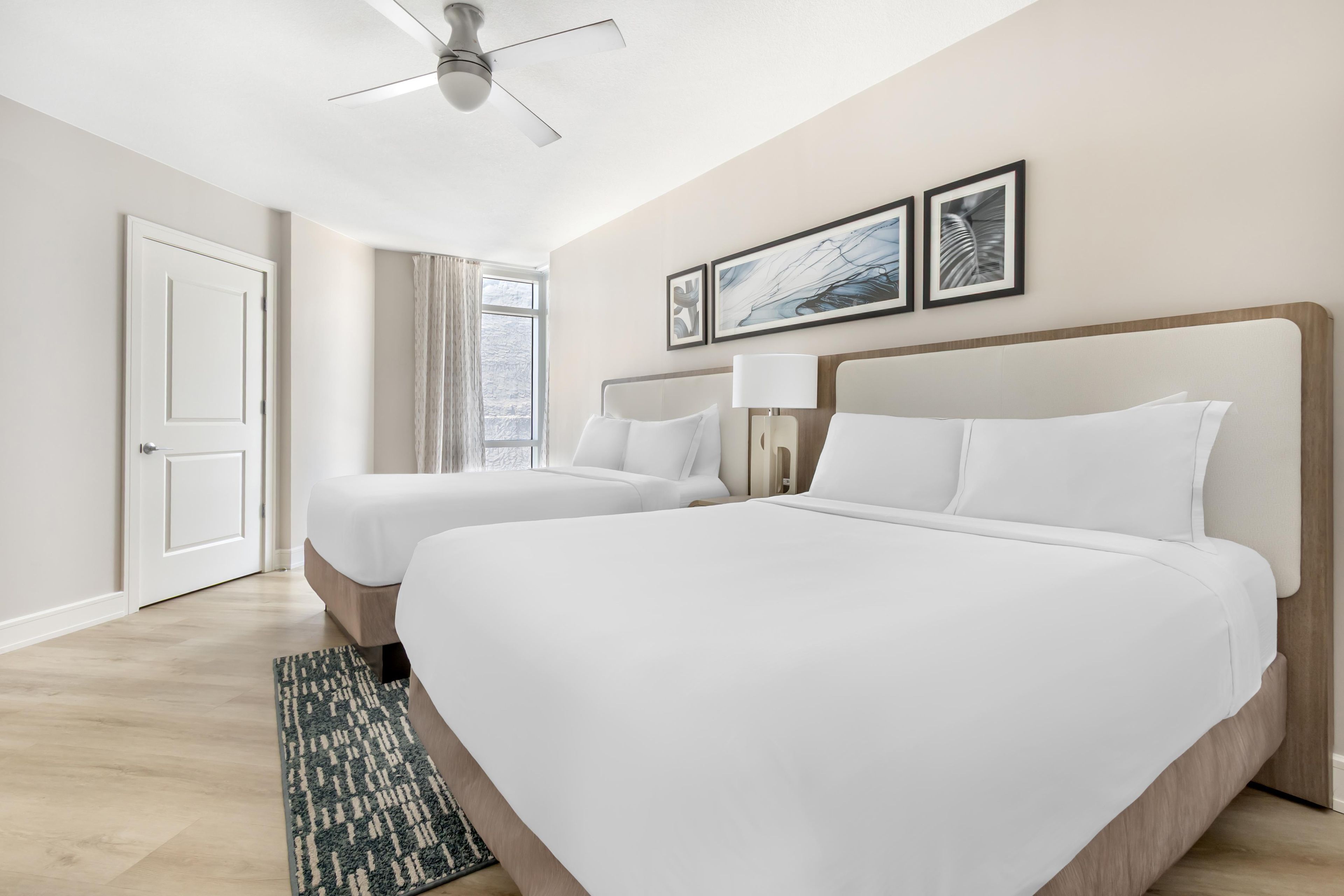 Ein Hotelzimmer mit zwei Betten, einem Deckenventilator, einem fensterbelaubten Fenster und gerahmten Kunstwerken an der Wand.