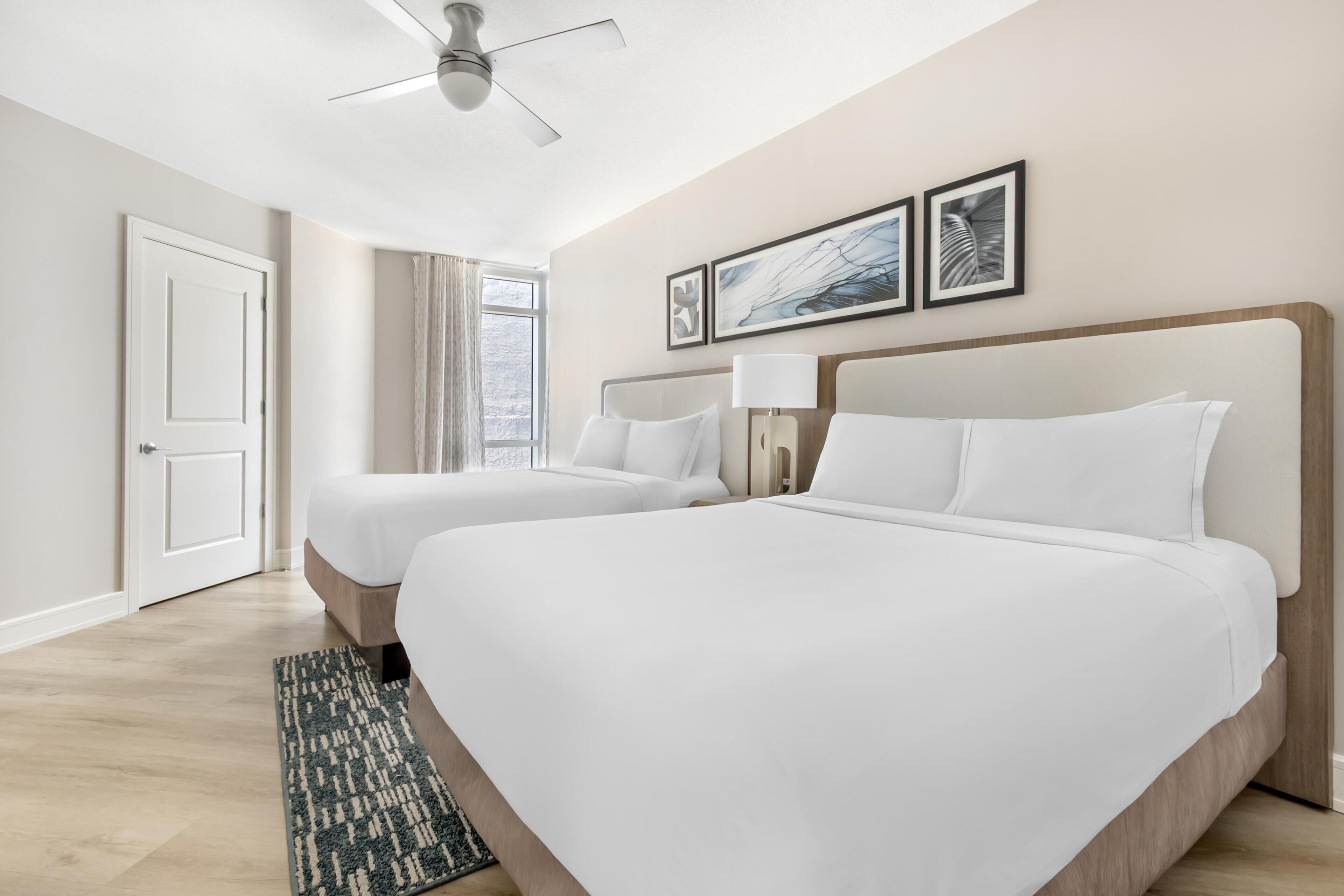 A hotel room with two beds, a ceiling fan, a window with curtains, and framed artwork on the wall.