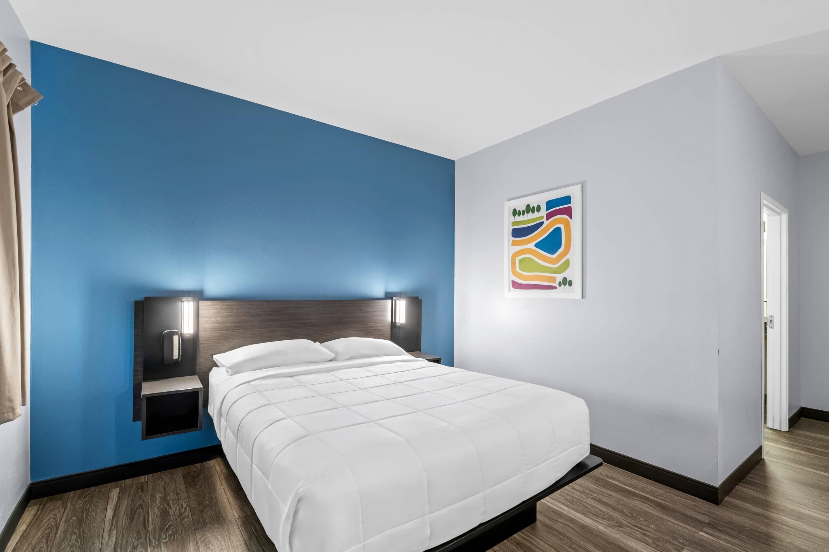 A modern hotel room with a large bed, a nightstand, a wall-mounted light, and a colorful abstract painting.
