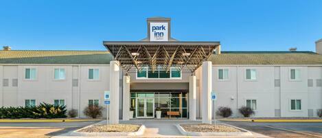 Entrada de un hotel Park Inn con un vestíbulo techado, estacionamiento para discapacitados y un cielo azul despejado.