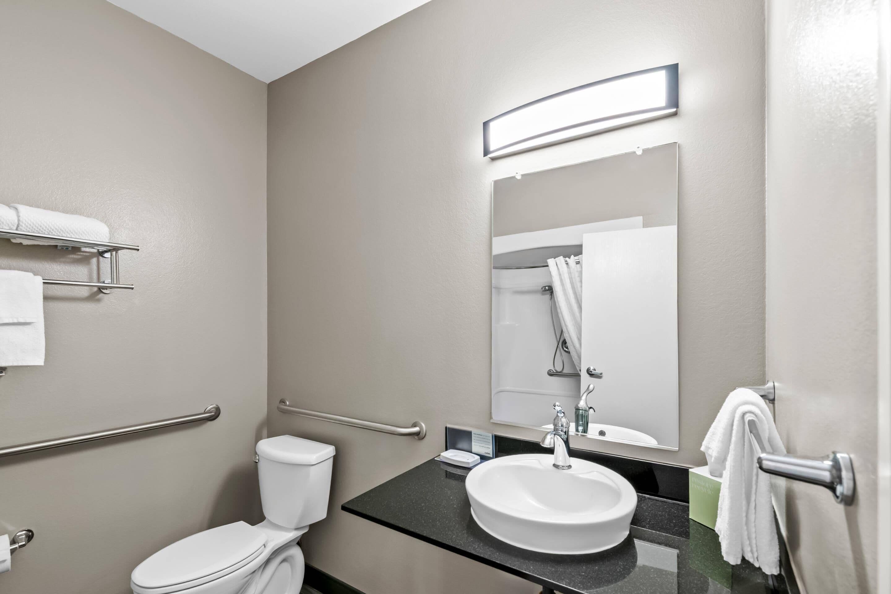 Une salle de bain équipée de toilettes, d’un lavabo, d’un miroir, d’un porte-serviettes et de barres d’appui.