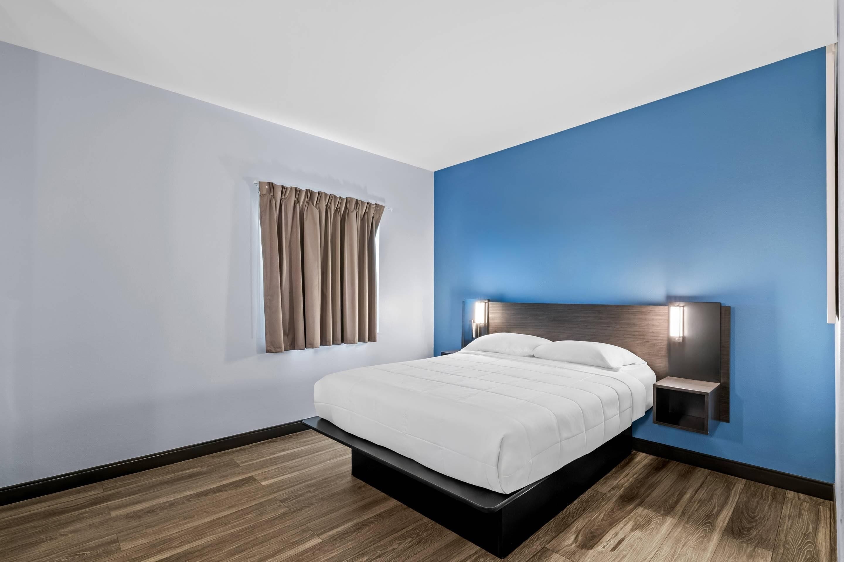 A modern hotel room with a bed, a nightstand, a wall-mounted light, and a window with curtains.