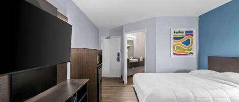 Une chambre d’hôtel dotée d’une grande télévision à écran plat, d’un bureau en bois, d’une chaise bleue, de deux lits, d’une salle de bain avec un miroir et d’une œuvre d’art abstraite et colorée accrochée au mur.