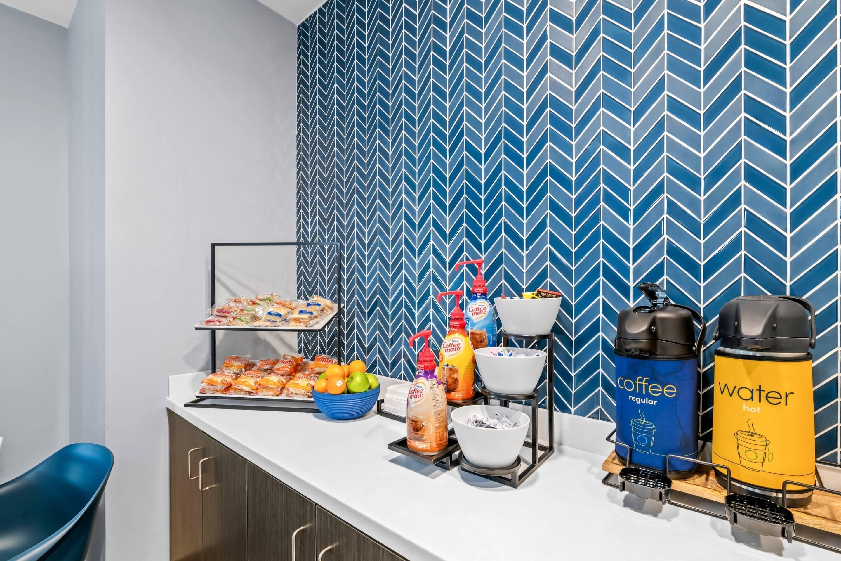 A breakfast bar with coffee and water dispensers, snacks, and a fruit bowl.
