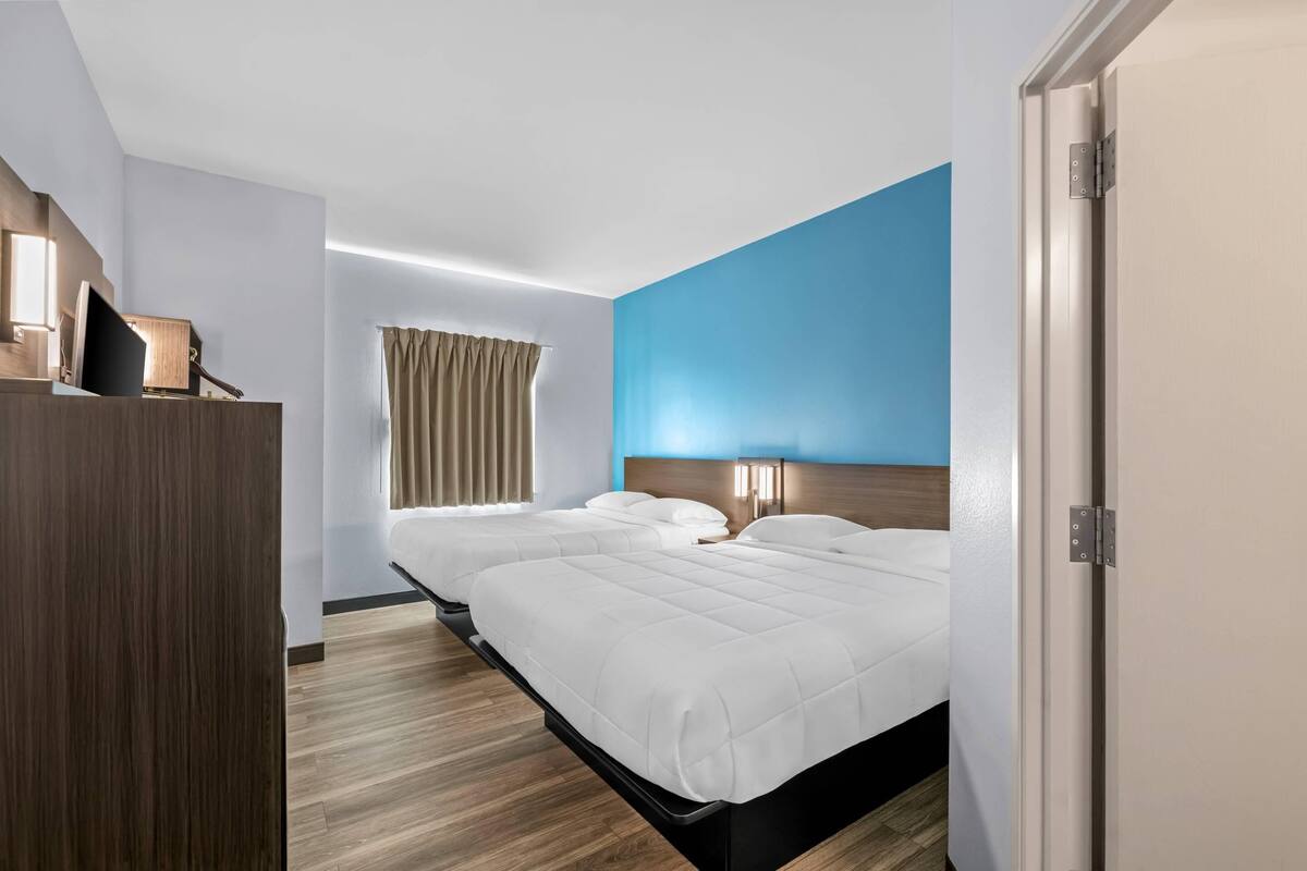 A hotel room with two beds, a wooden wardrobe, a bedside table with a lamp, and a window with curtains.