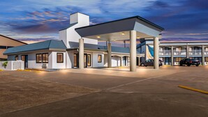 Una estación de servicio moderna con zona de surtidor techada, un pequeño edificio de techo azul y un estacionamiento con varios coches.