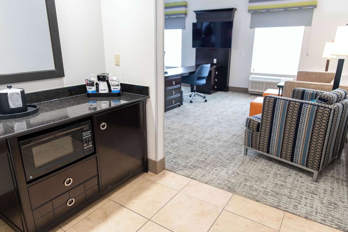 A hotel room with a kitchenette, a desk, a chair, a sofa, and a window.