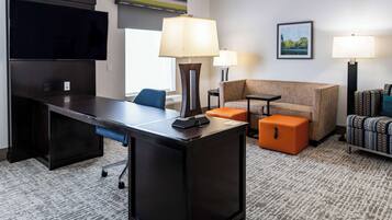 A hotel room with a flat-screen TV, a desk with a lamp, a sofa, an orange ottoman, and a patterned armchair.