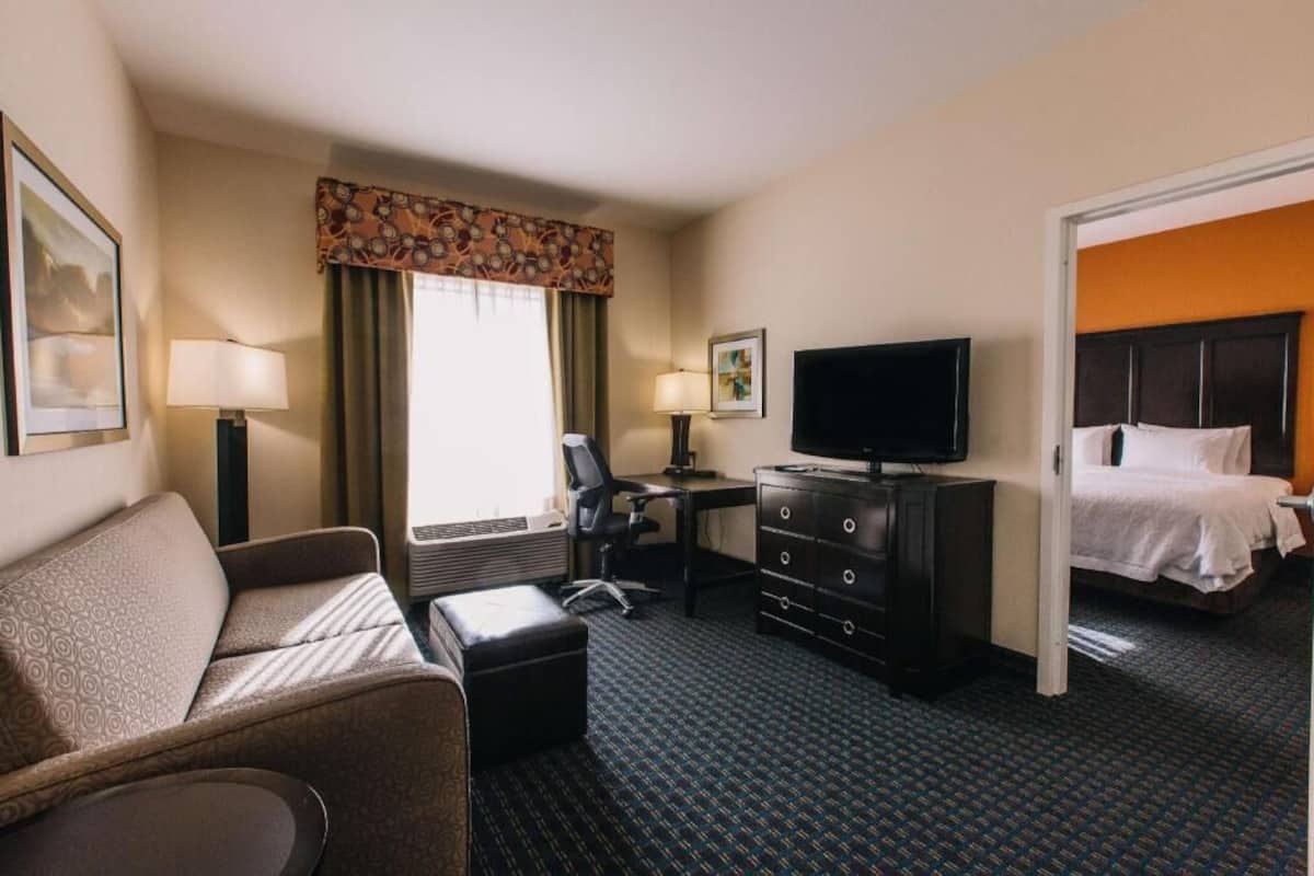 A hotel room with a sofa, a desk with a computer, a TV, a bed, and a window with curtains.