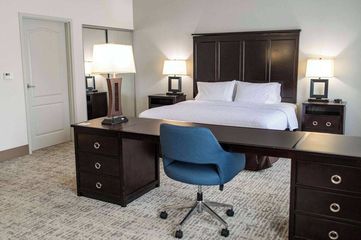 A hotel room with a large bed, two bedside tables with lamps, a desk with a chair, and a door.