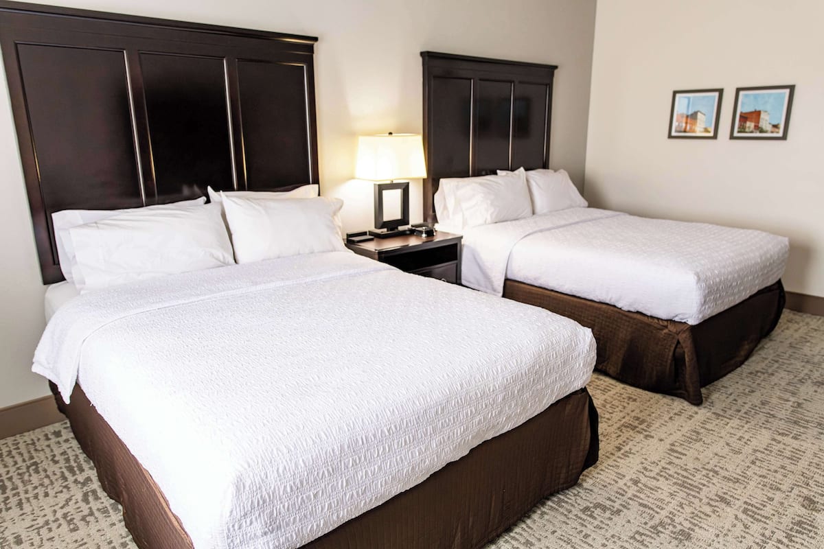 A hotel room with two beds, dark wood headboards, a nightstand with a lamp, and a carpeted floor.