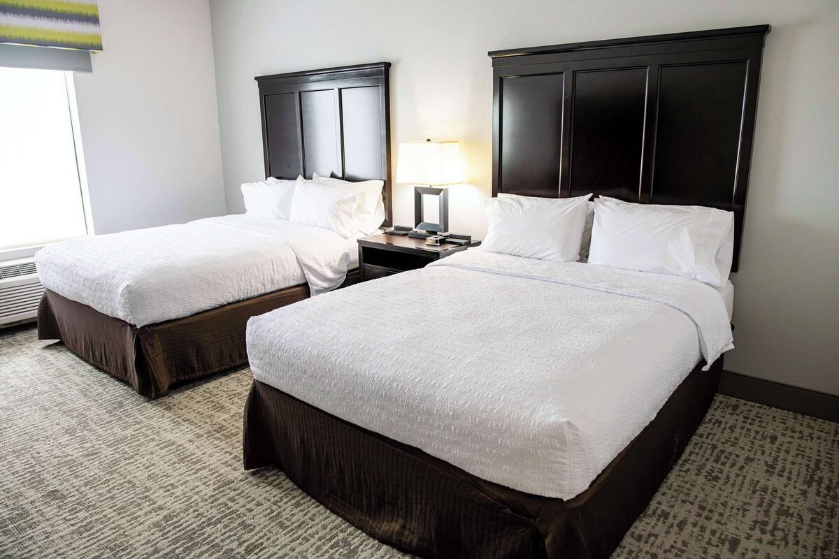 A hotel room with two beds, dark headboards, a nightstand with a lamp, and a window with blinds.