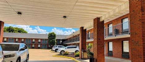 Un estacionamiento con varios coches, un corredor cubierto y un edificio de paredes de ladrillo y ventanas.