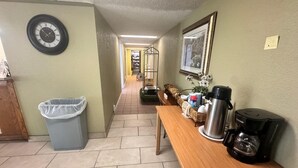 A hallway with a trash bin, a wooden table with a coffee maker, a framed picture, and a clock on the wall. - Delta Inn West Helena US 49 AR (West Helena)