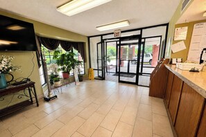 A reception area with a flat-screen TV, a reception desk, potted plants, and an exit door. - Delta Inn West Helena US 49 AR (West Helena)