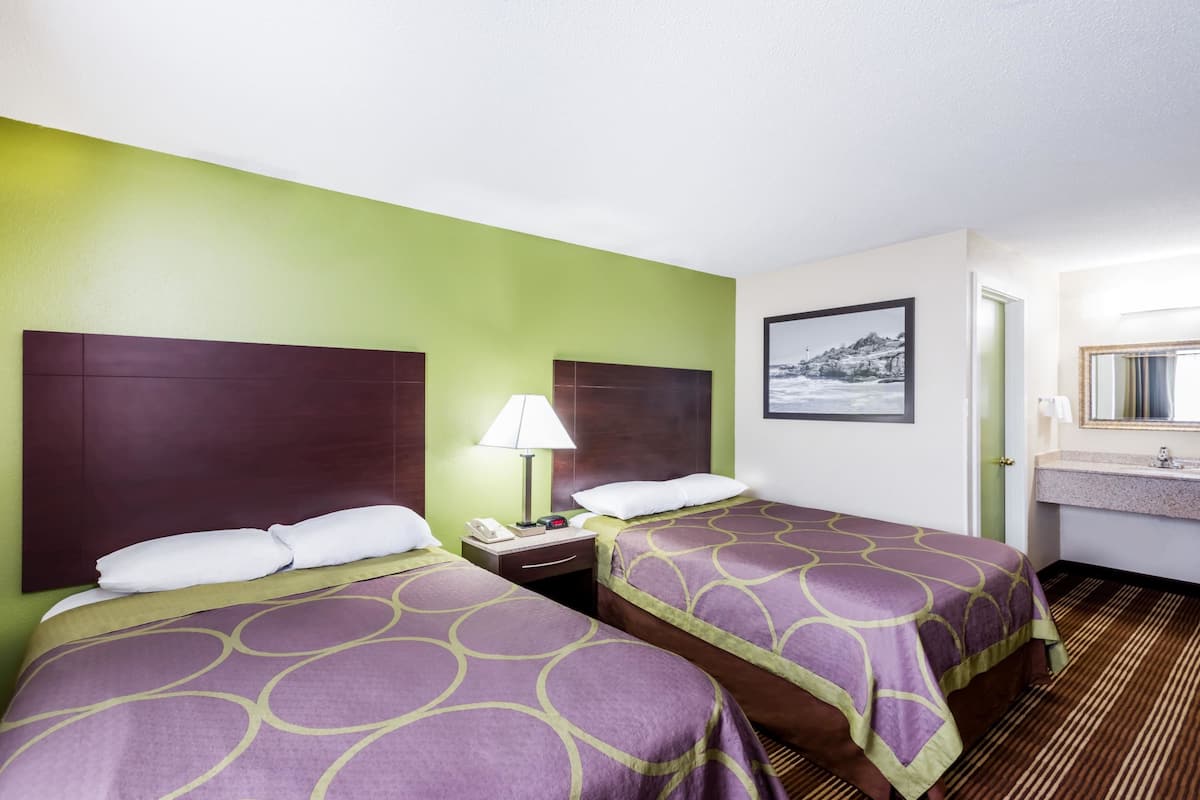 Ein Hotelzimmer mit zwei Betten, einem Nachttisch mit Lampe, einem gerahmten Bild an der Wand und einem Badezimmer mit Waschbecken und Spiegel.