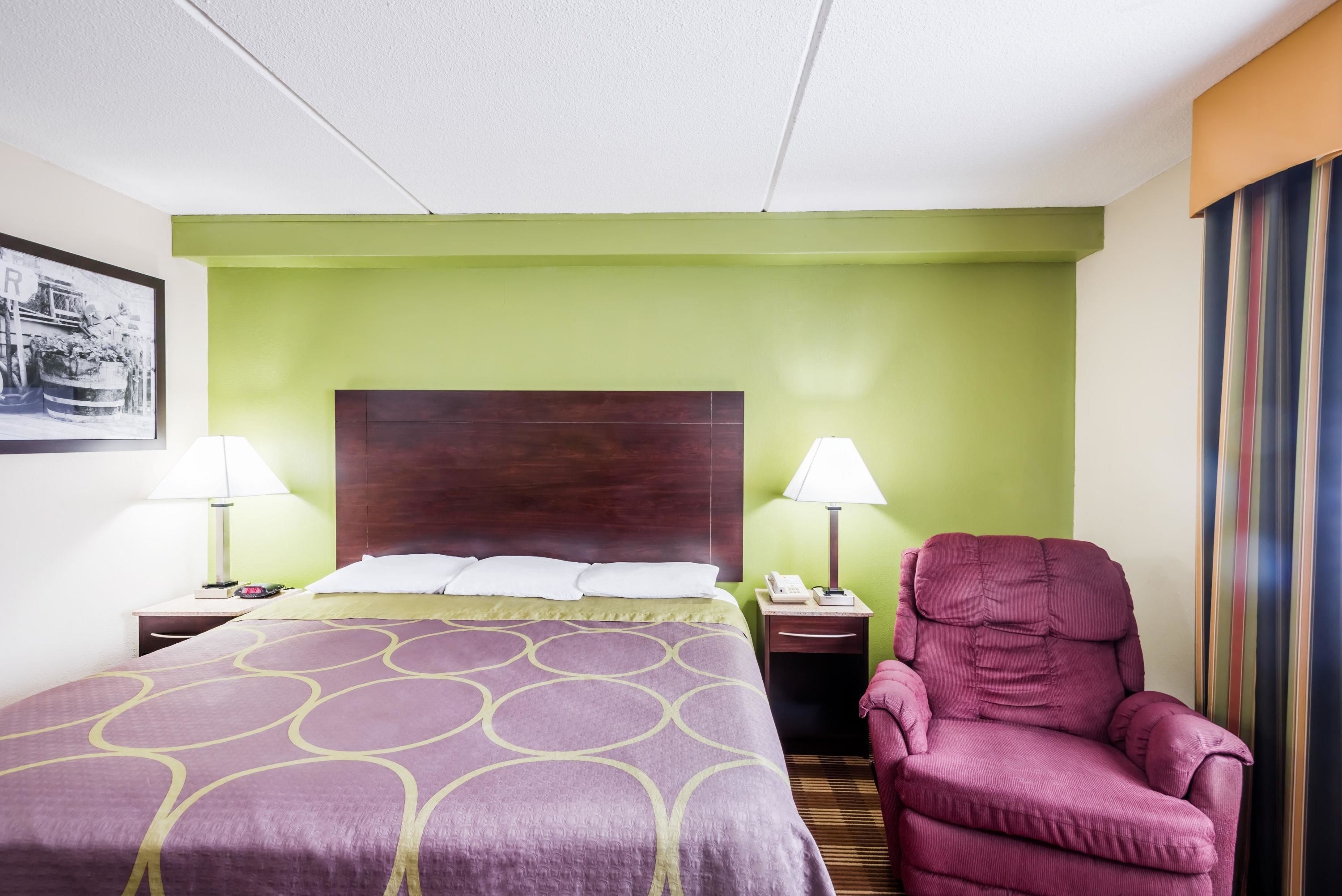 A hotel room with a large bed, two bedside tables with lamps, a wooden headboard, and a single purple armchair.