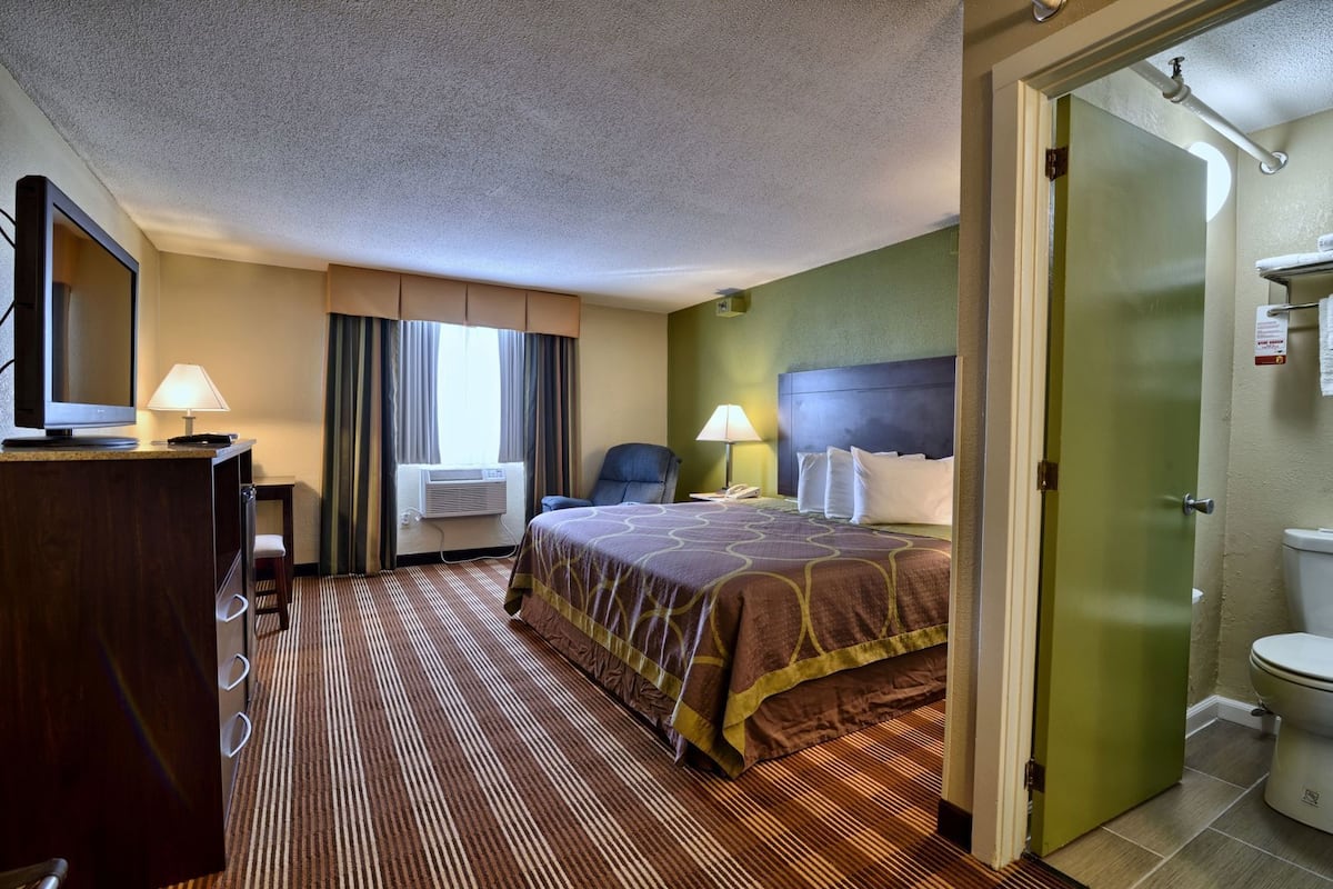 Ein Hotelzimmer mit einem Bett, einem Schreibtisch mit Lampe, einer Klimaanlage, einem Stuhl und einem Badezimmer mit Toilette und Dusche.