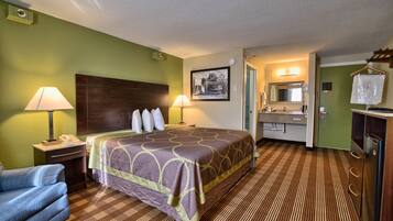 A hotel room with a bed, two bedside lamps, a nightstand, a chair, a bathroom with a sink and mirror, and a refrigerator.