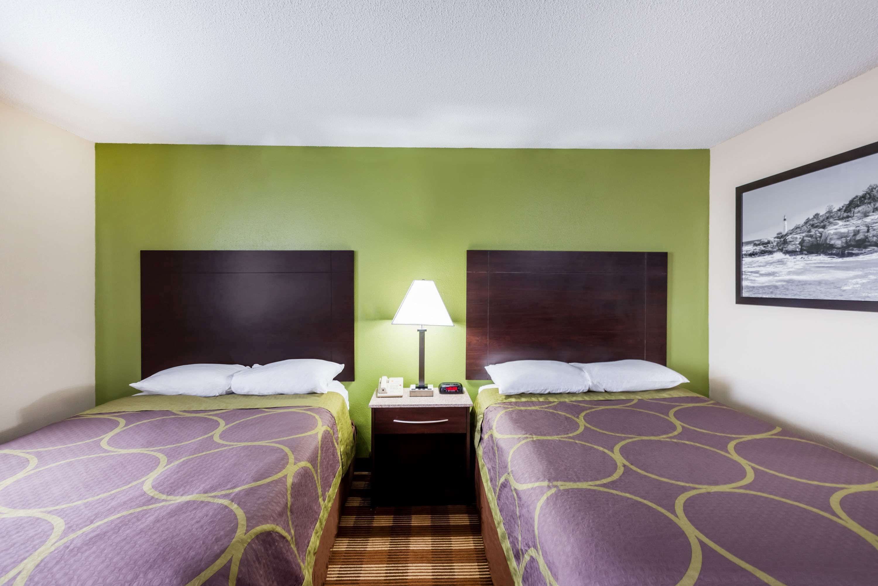 Habitación de hotel con dos camas, una mesita de noche con lámpara y un cuadro en la pared.