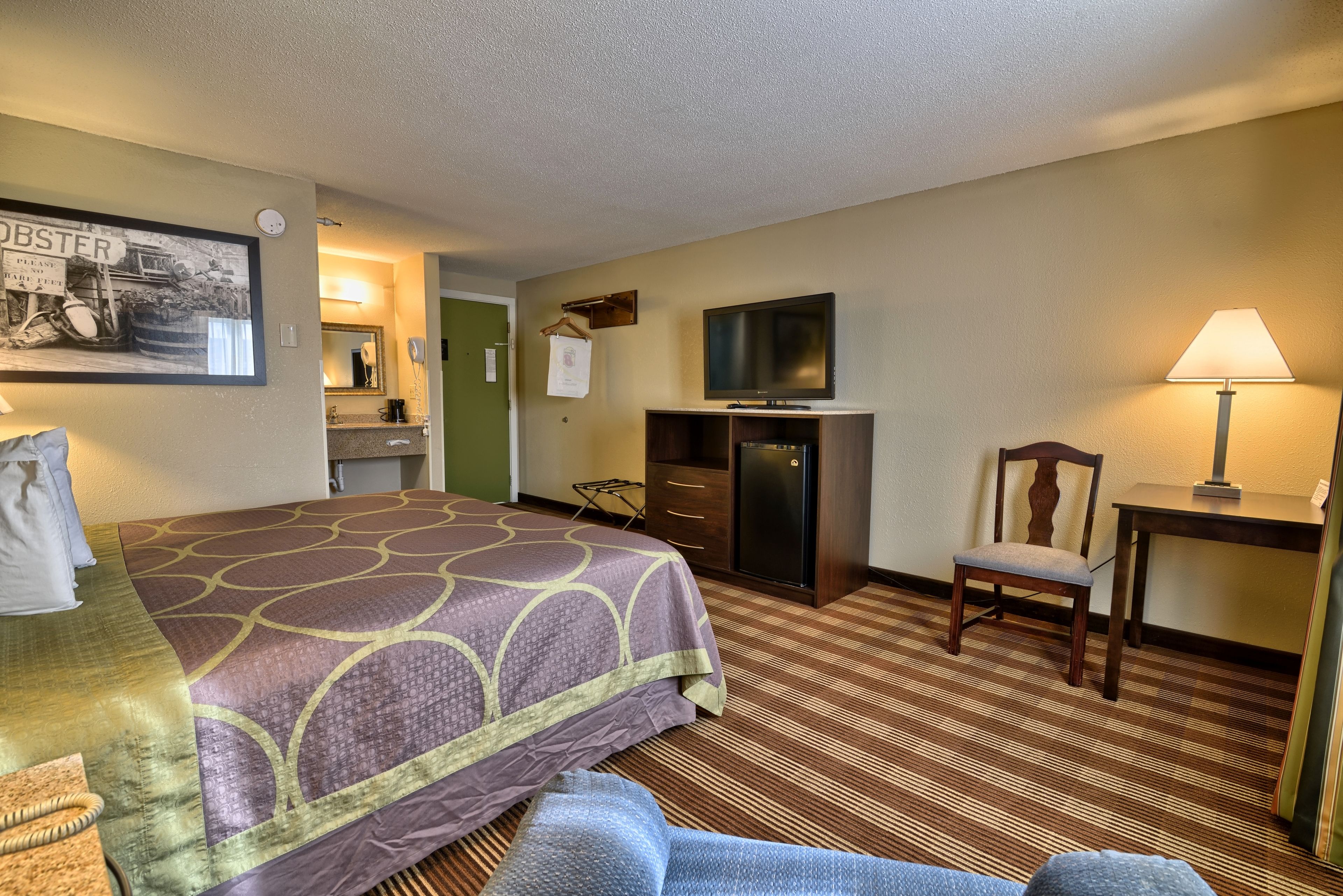 A hotel room with a large bed, a television, a desk with a lamp, a chair, and a dresser.