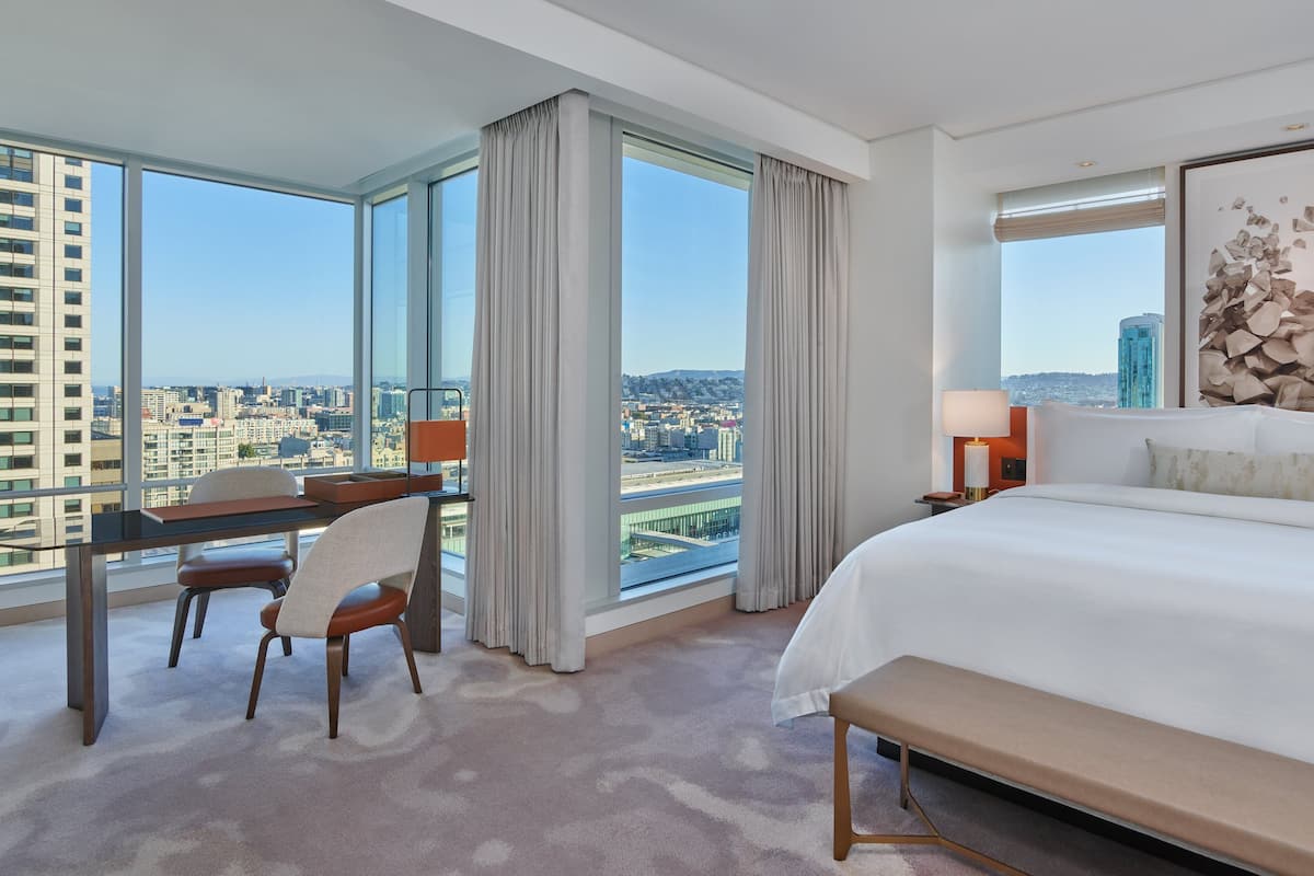 A hotel room with a large bed, a desk with a chair, a bench, and a view of the city through large windows.