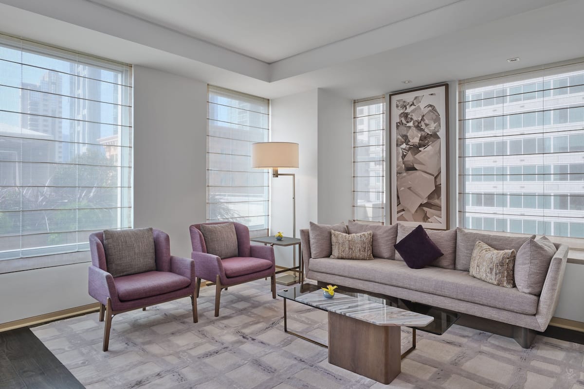 A modern living room with a sofa, two armchairs, a coffee table, and a floor lamp. Large windows provide natural light and views of the city.