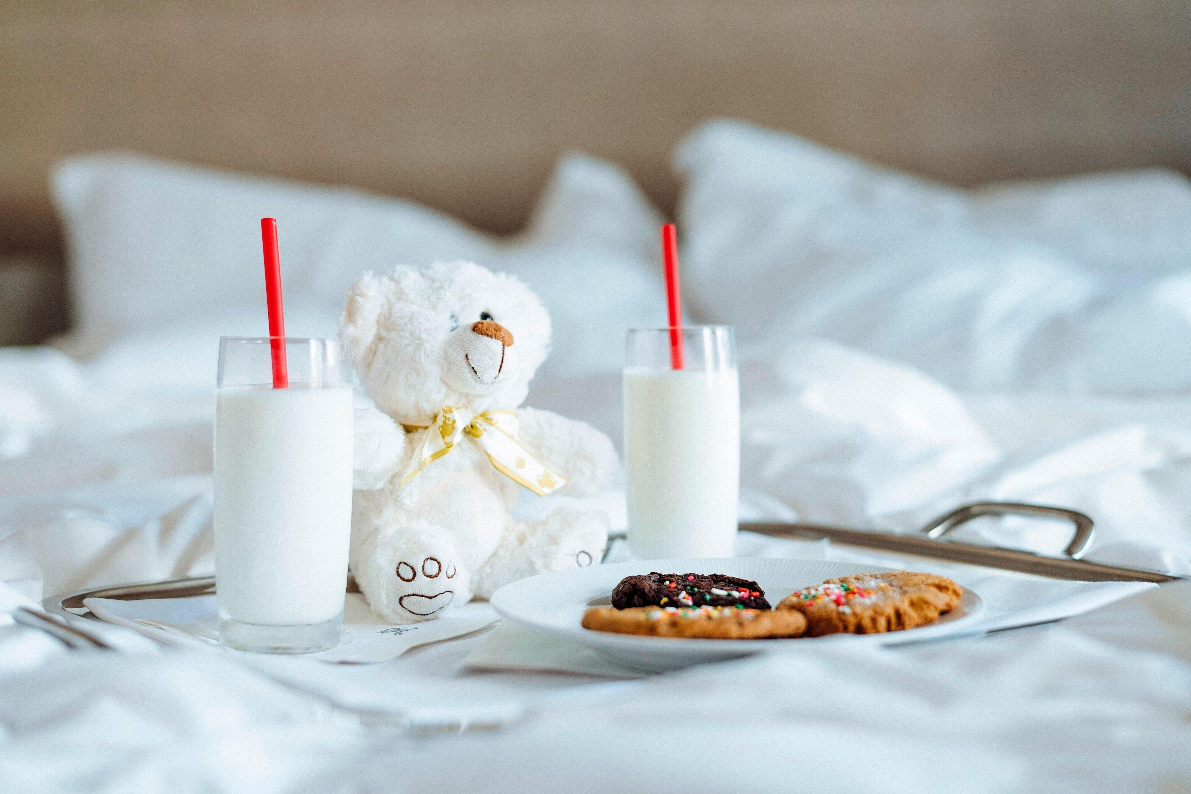 Una cama con un oso de peluche, dos vasos de leche y galletas.