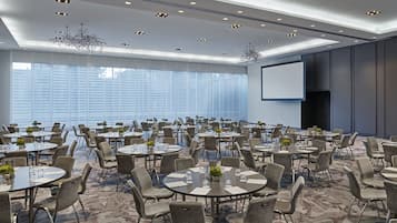 Un amplio salón de conferencias con mesas y sillas redondas, una pantalla de proyector y un ventanal con persianas.