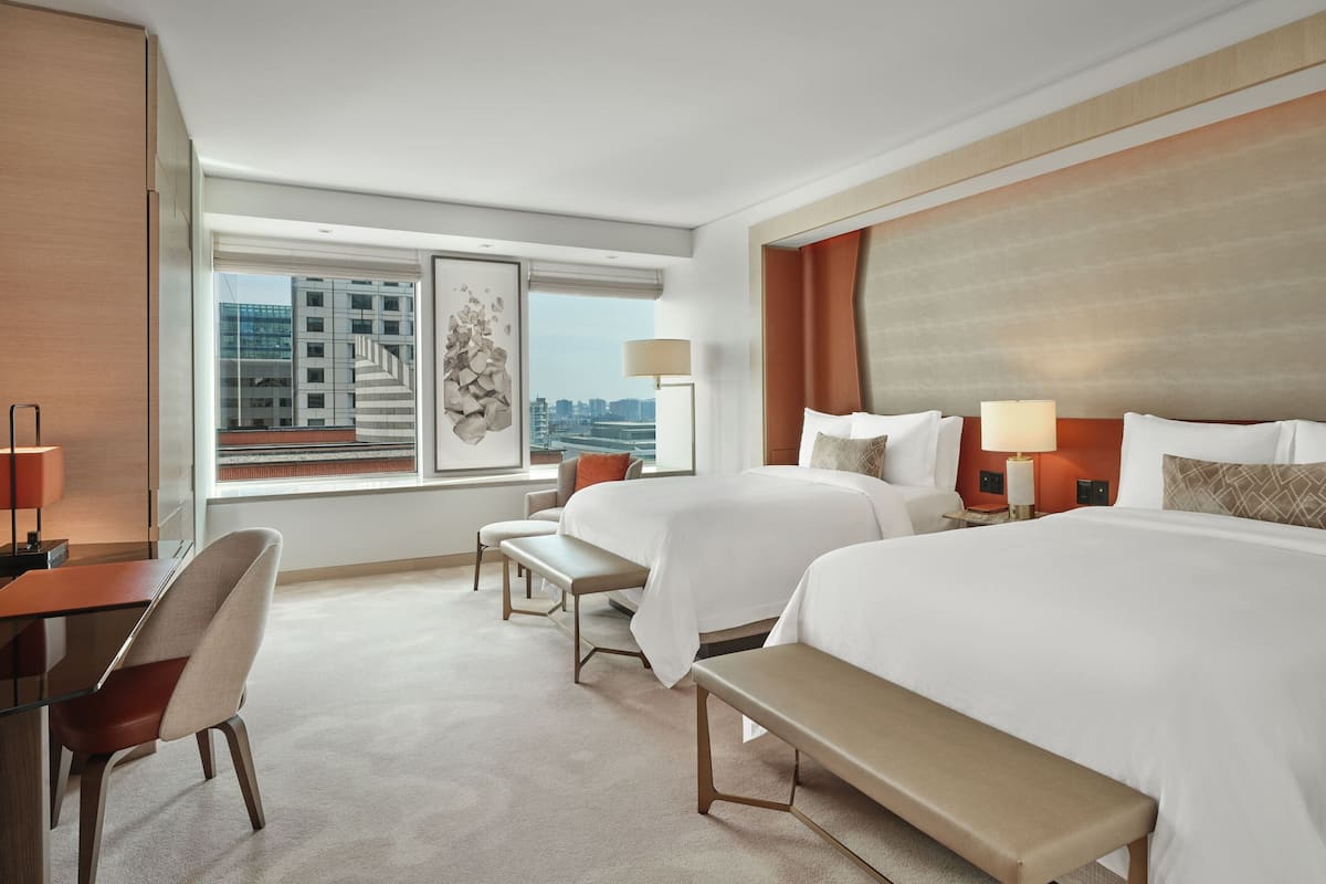 A hotel room with two beds, a desk, a chair, a bench, and a large window offering a city view.