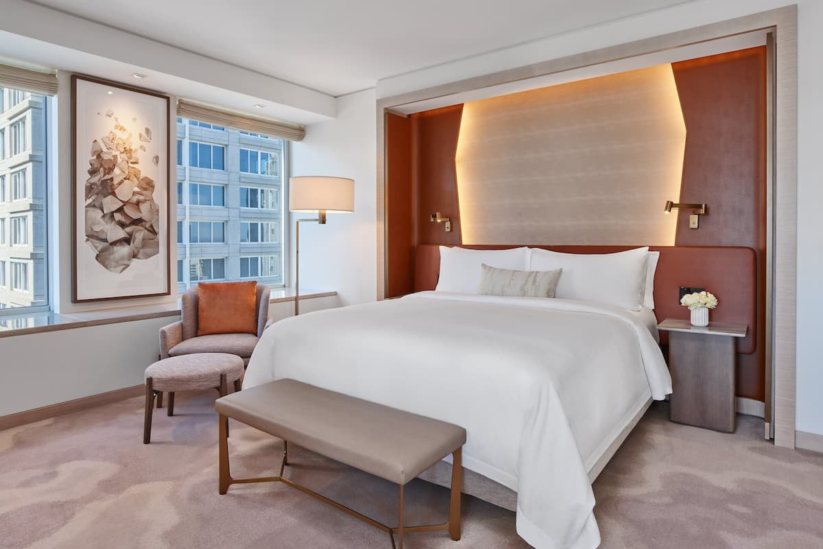 A modern hotel room with a large bed, a bench, a chair, a bedside table with a vase of flowers, and a framed artwork on the wall.