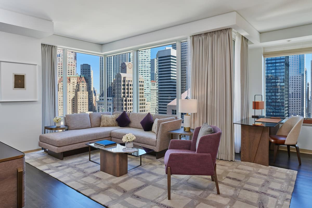 A modern hotel room with a large window offering a city view, a sofa, a chair, a coffee table, and a desk with a lamp.