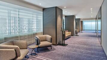 A modern hotel lobby with a carpeted floor, seating area with sofas and a small table, and large windows offering a view of the cityscape.
