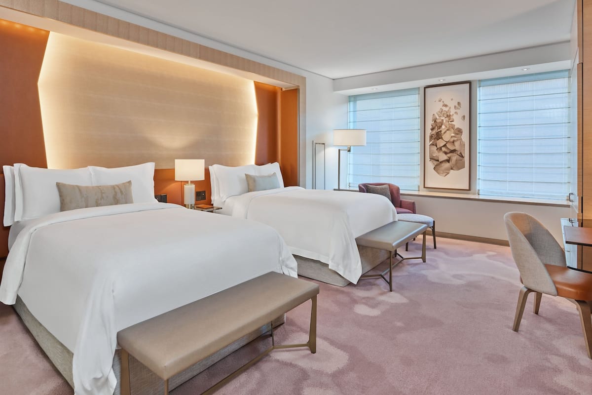 A hotel room with two beds, a bench, a desk, a chair, and a large window with blinds.