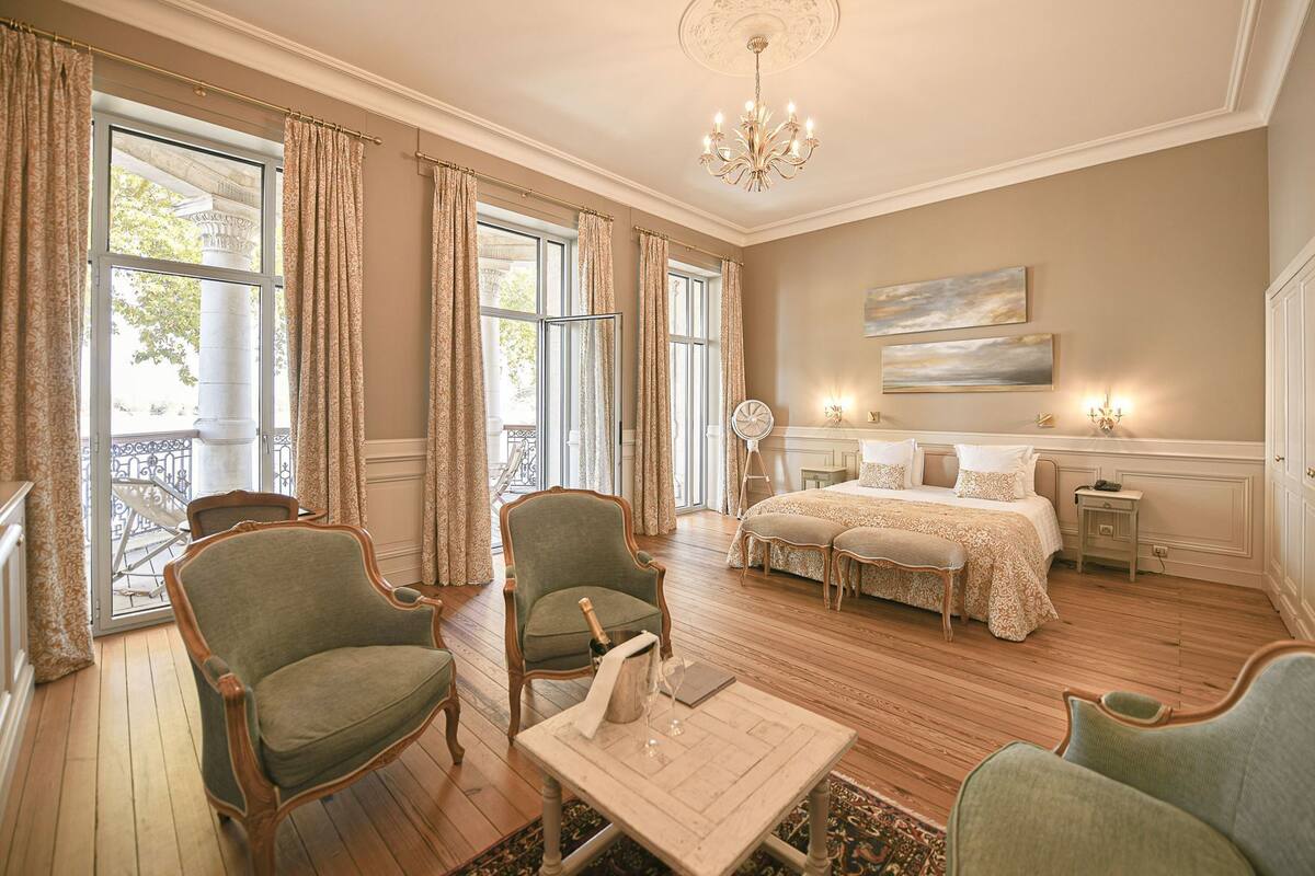 A bedroom with a large bed, a sitting area with chairs, a table with a wine glass, and a balcony with curtains.