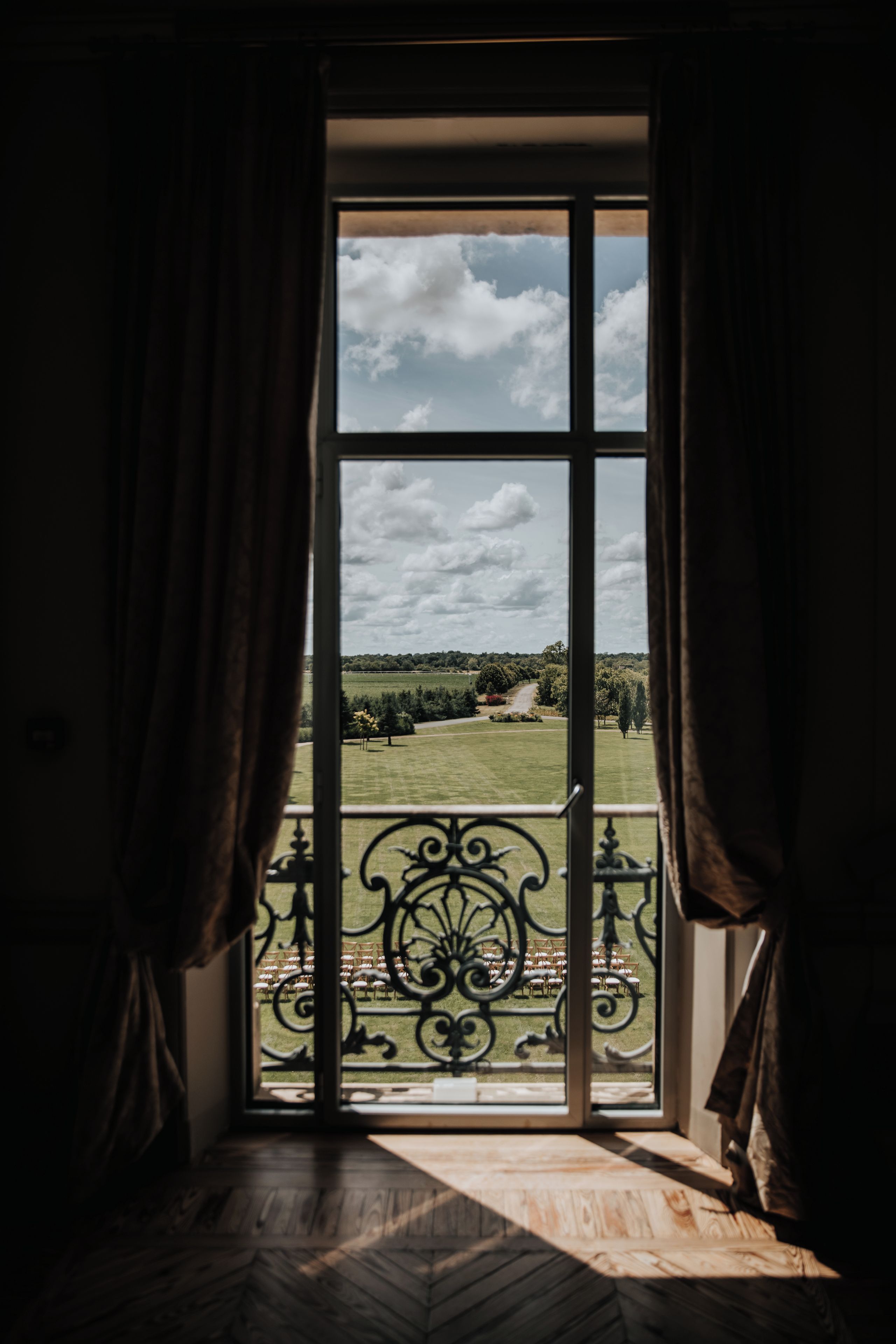 A room with a large window, curtains, and a view of a green landscape with trees and a path.
