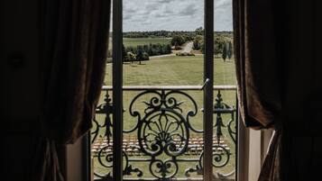 A room with a large window, curtains, and a view of a green landscape with trees and a path.