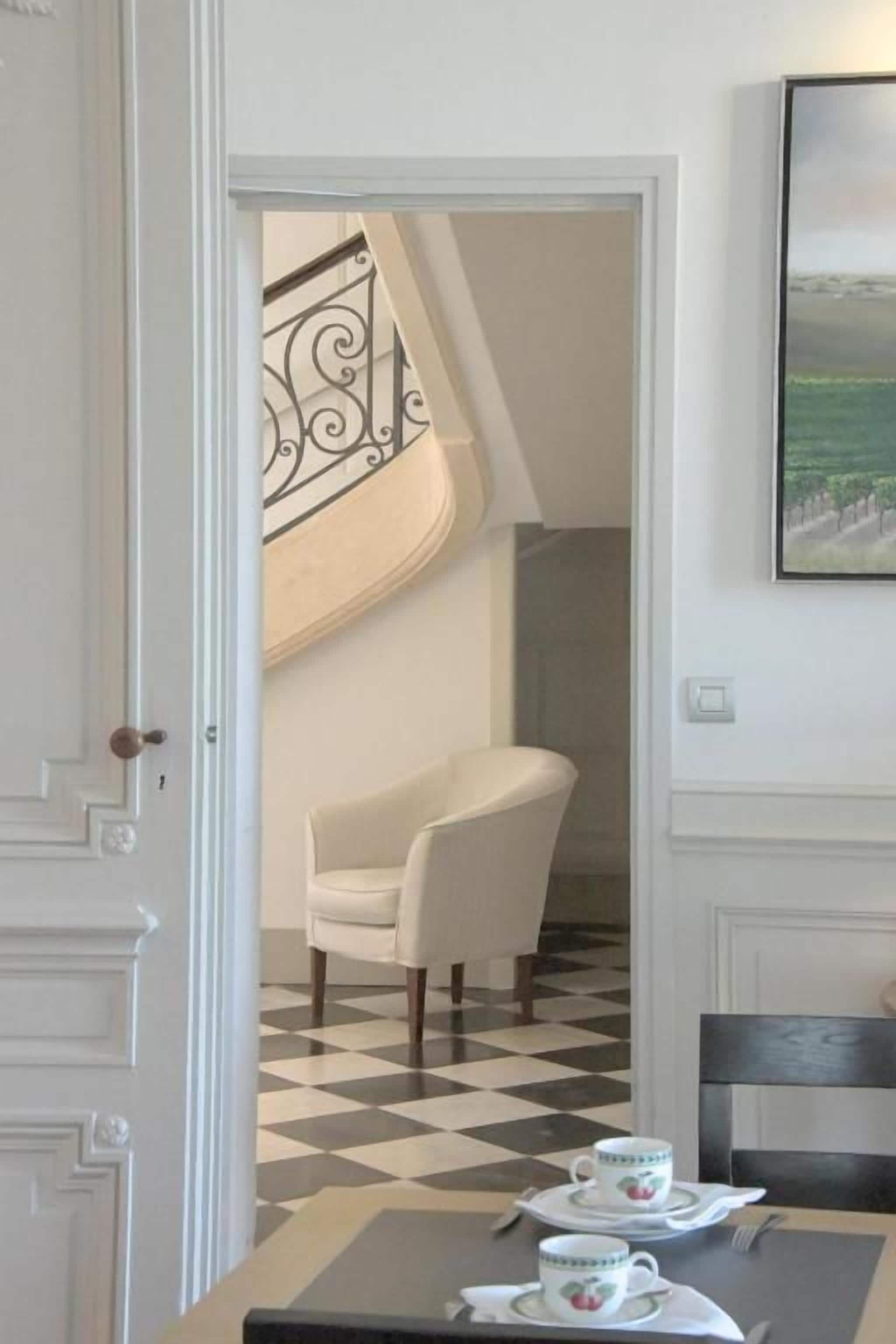 Une pièce avec un sol à carreaux, une table à manger avec des tasses à thé, une chaise blanche et un escalier orné d’une rampe décorative.