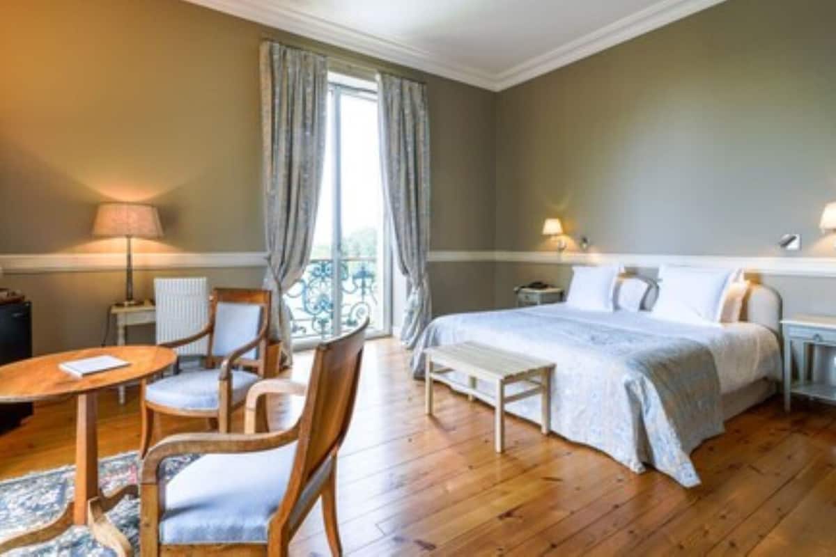 A hotel room with a bed, bedside tables, a chair, a small table, and a window with curtains.