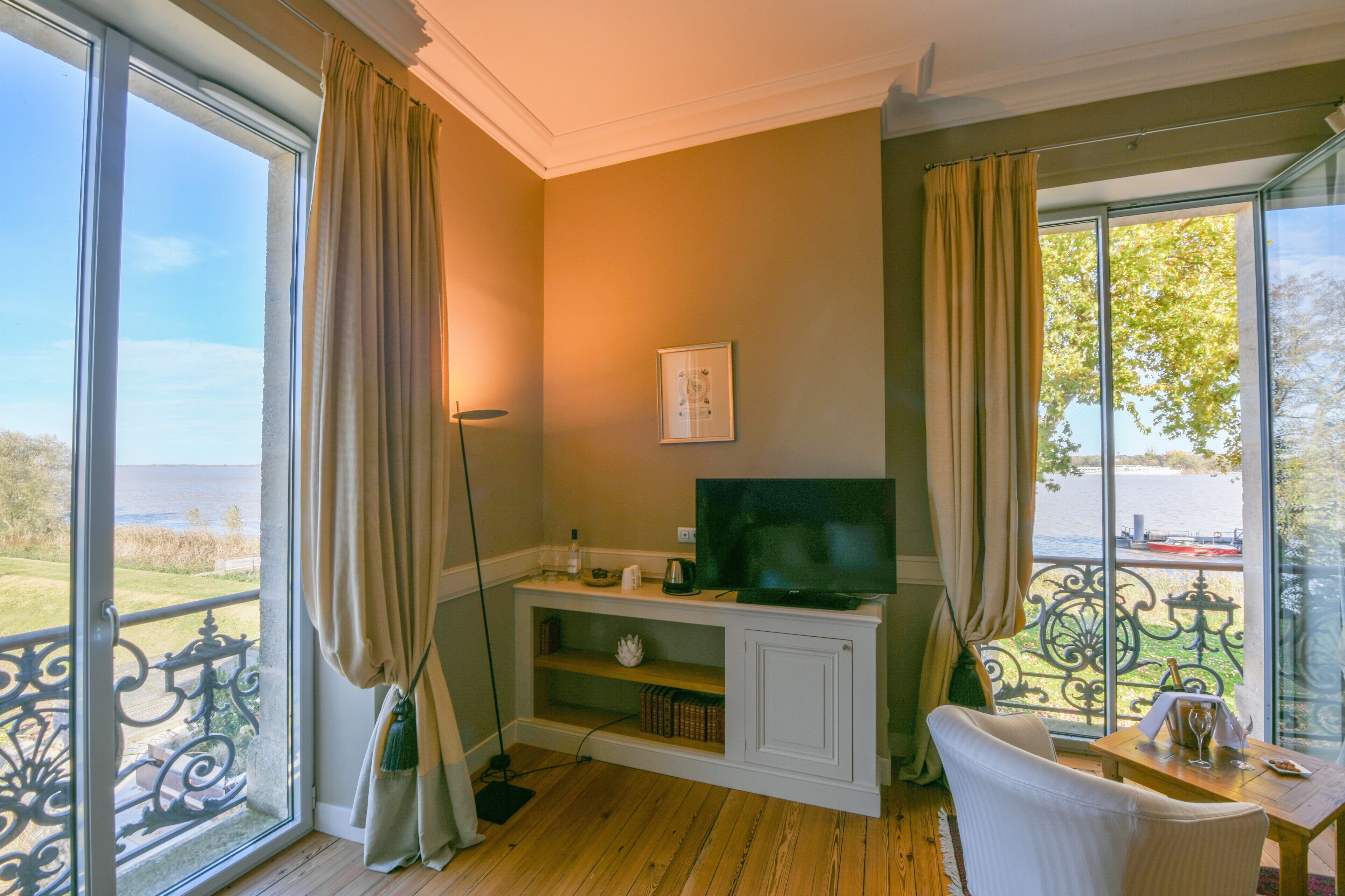 A room with a TV on a wooden cabinet, a chair, a table with a glass, and a view of the outdoors.