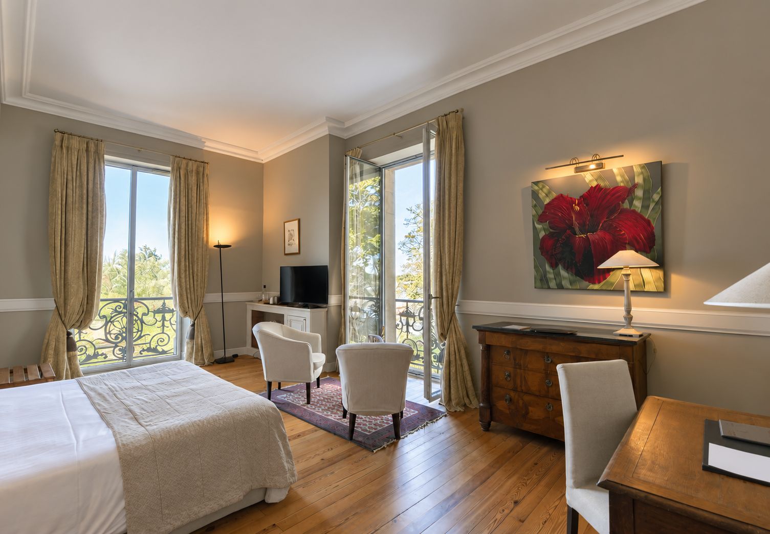 Quarto de hotel com cama, duas cadeiras, escrivaninha, televisão e uma janela grande com cortinas.
