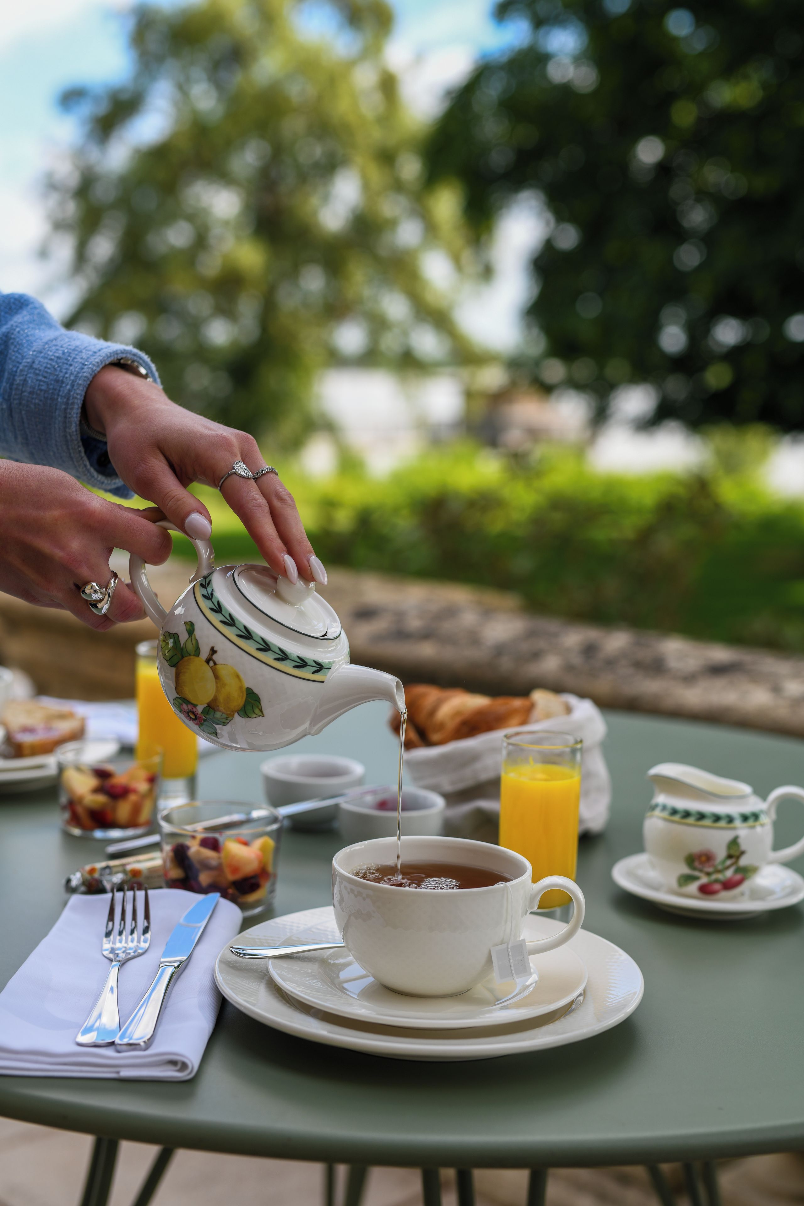A table set for breakfast with a teapot pouring tea into a cup, a glass of orange juice, and a croissant.
