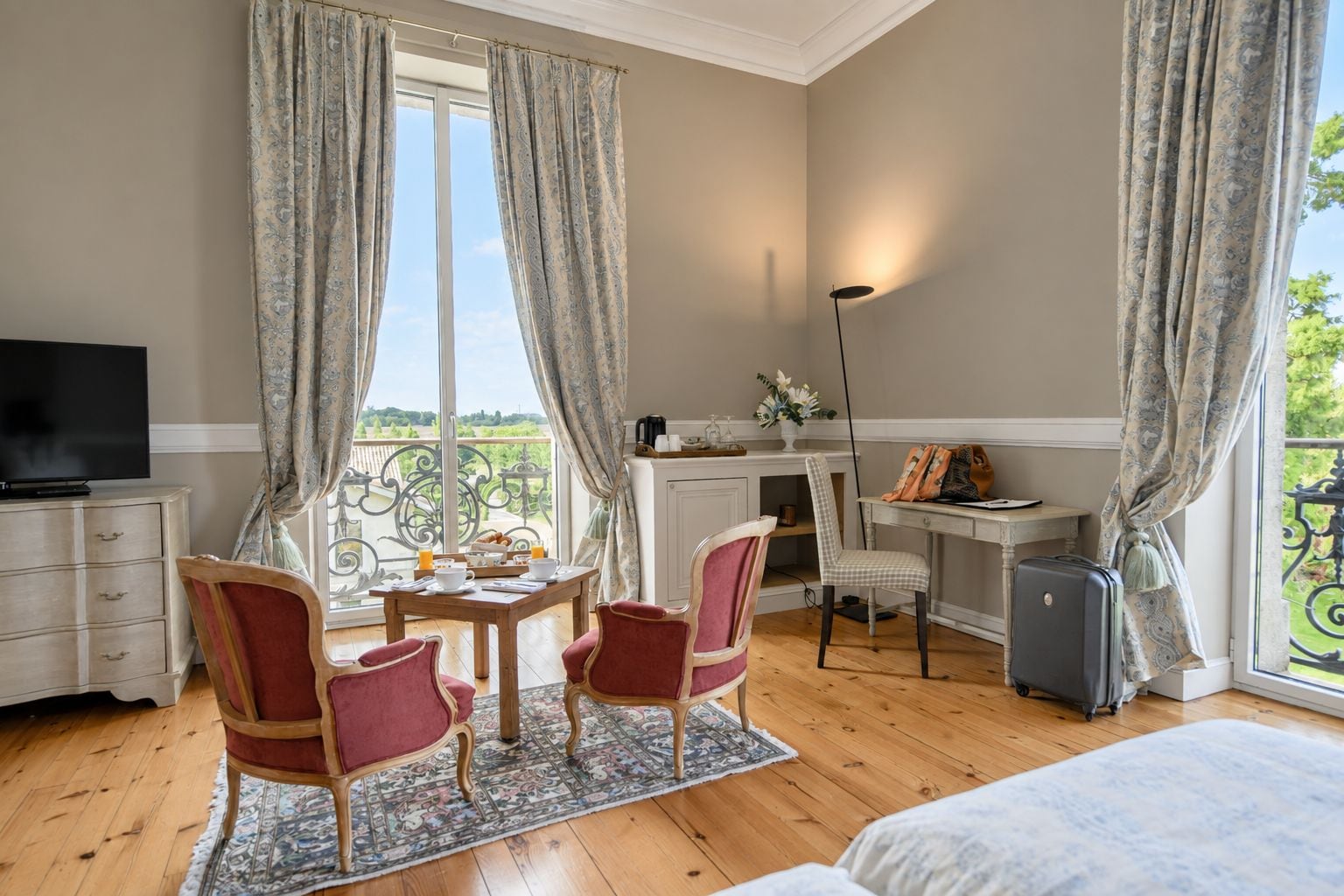 A hotel room with a bed, a desk, a television, a chair, a table with breakfast items, and a view of the outdoors.