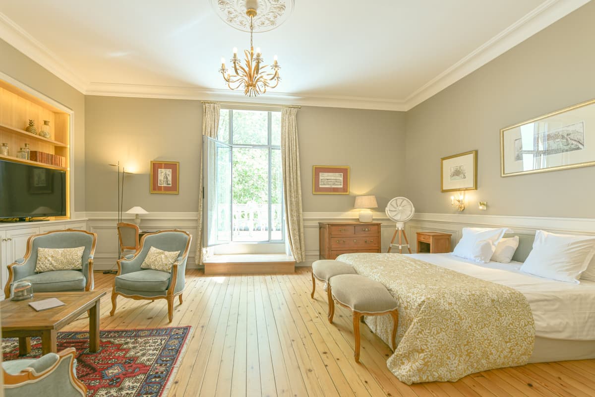 A bedroom with a large bed, two armchairs, a wooden floor, a chandelier, and a window with curtains.