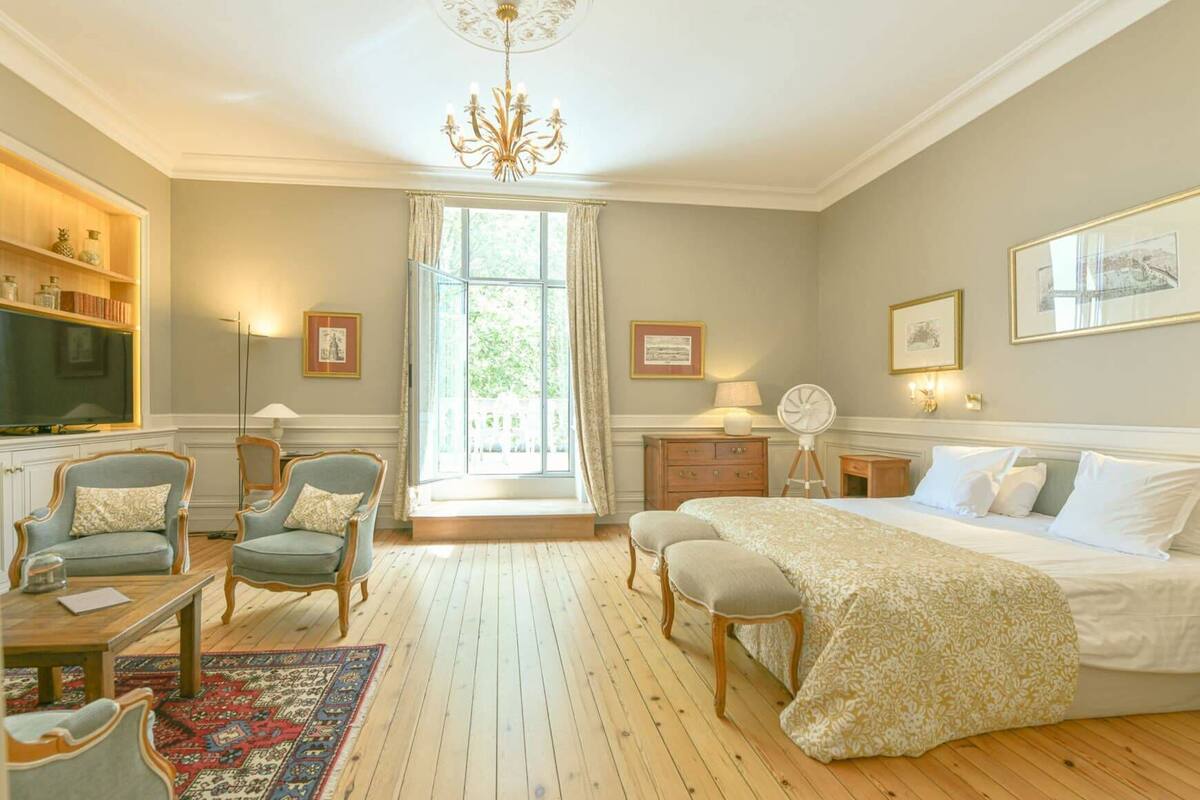 A hotel room with a large bed, two armchairs, a wooden floor, a chandelier, and a window with curtains.